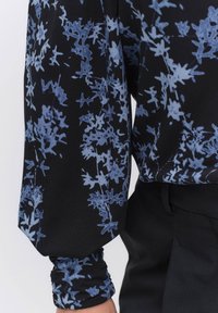 Svart blus med ett blommönster i olika nyanser av blått. Mjuk tyg med lätt puffade ärmar och samlade manschetter.