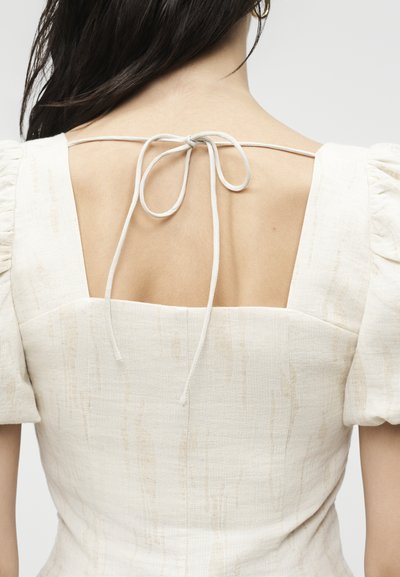 Πίσω όψη ατόμου που φοράει κρεμ φόρεμα με τετράγωνη λαιμόκοψη, φουσκωτά μανίκια και λεπτό ιμάντα δεμένο φιόγκο στο πάνω μέρος της πλάτης.