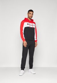 Sudadera con cremallera en rojo y negro, con un panel blanco y el logo de "Champion", combinada con pantalones de chándal negros y zapatillas blancas, todo sobre un fondo sencillo.