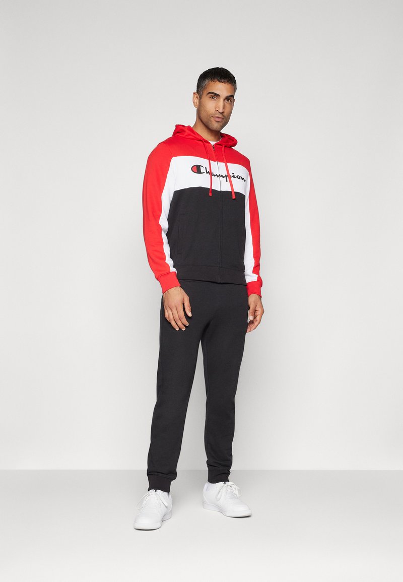 Sudadera con cremallera en rojo y negro, con un panel blanco y el logo de "Champion", combinada con pantalones de chándal negros y zapatillas blancas, todo sobre un fondo sencillo.