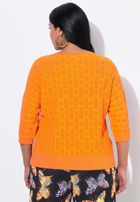 Oranger Strickpullover mit einem strukturierten Muster, 3/4-Ärmeln und einem gerippten Saum. Kombiniert mit schwarzen Hosen, die mit einem bunten Schmetterlingsdruck verziert sind.