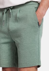 Green shorts made of textured fabric with a drawstring waistband and side pockets. Features a branded tag on the side.