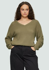 Olijfgroen gebreide trui met een V-hals, lange mouwen en geribbelde manchetten, gecombineerd met zwarte broek. Gelaagd stof met een ontspannen pasvorm.