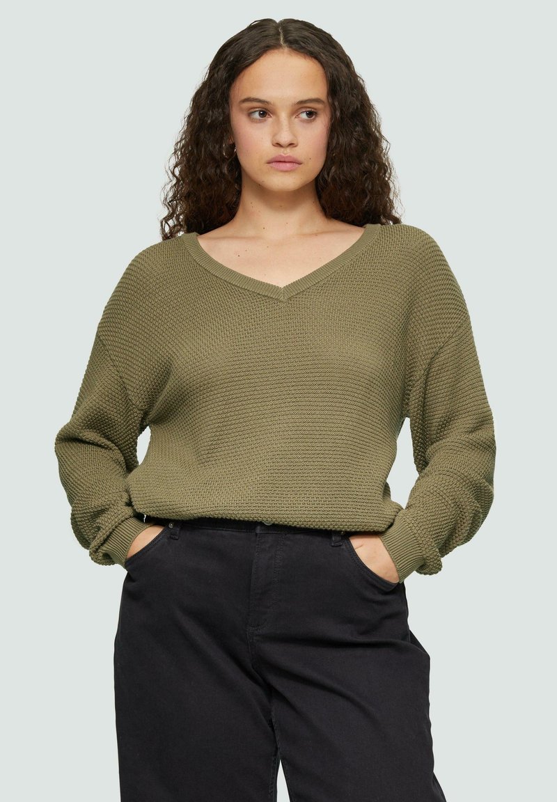 Olijfgroen gebreide trui met een V-hals, lange mouwen en geribbelde manchetten, gecombineerd met zwarte broek. Gelaagd stof met een ontspannen pasvorm.