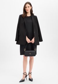 Zwarte blazer met brede revers, gedragen over een zwarte jurk. Geaccentueerd met een zwarte handtas met gouden details en stilettohakken.