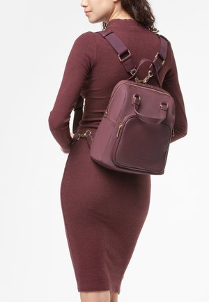 Donna che indossa un abito lungo aderente di colore bordeaux, mentre porta uno zaino in pelle bordeaux abbinato con cerniere dorate e spalline regolabili.