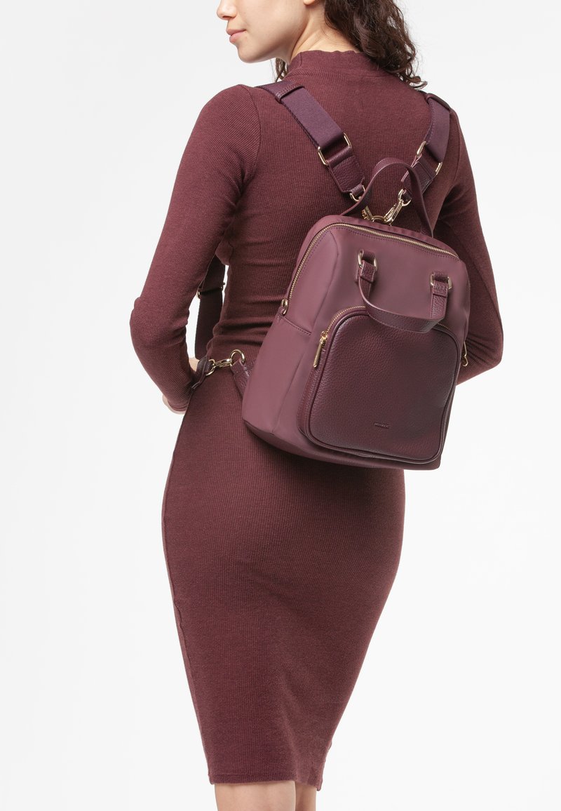 Donna che indossa un abito lungo aderente di colore bordeaux, mentre porta uno zaino in pelle bordeaux abbinato con cerniere dorate e spalline regolabili.