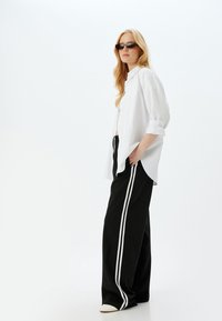 Camisa blanca de botonadura con mangas remangadas, combinada con pantalones negros holgados que tienen franjas laterales blancas. Los accesorios incluyen gafas de sol oscuras.