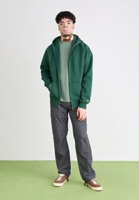 Grüne Kapuzenjacke mit Reißverschluss, graue Cargo-Hosen und braune Sneakers. Die Jacke hat Fronttaschen und minimalistische Hardware-Details. Glatte Textur.