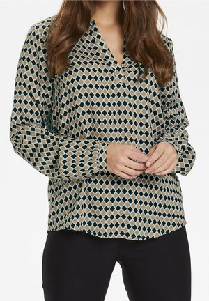 Femme portant un chemisier beige à manches longues avec un motif losanges noir et vert, debout, les mains jointes devant, associée à un pantalon noir.