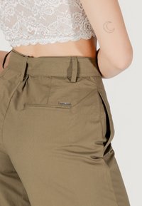 Pantaloni khaki a vita alta con tasca laterale, caratterizzati da una superficie morbida e passanti per cintura. Il tessuto appare leggero e versatile.