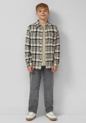 Chłopiec stojący oparty o ścianę, ubrany w beżową koszulę, kraciastą koszulę na guziki, szare dżinsy i białe trampki, lekko się uśmiechając.