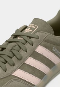 Κοντινό πλάνο του αθλητικού παπουτσιού Adidas Gazelle σε ελαιί πράσινο χρώμα, με σουέτ υλικό, ροζ ρίγες και κορδόνια στο ίδιο πράσινο χρώμα.
