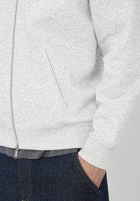 Haut de sweat à capuche zippé gris clair avec une finition texturée, orné d'une poche latérale et de poignets côtelés, superposé à une chemise gris foncé et à un jean en denim.