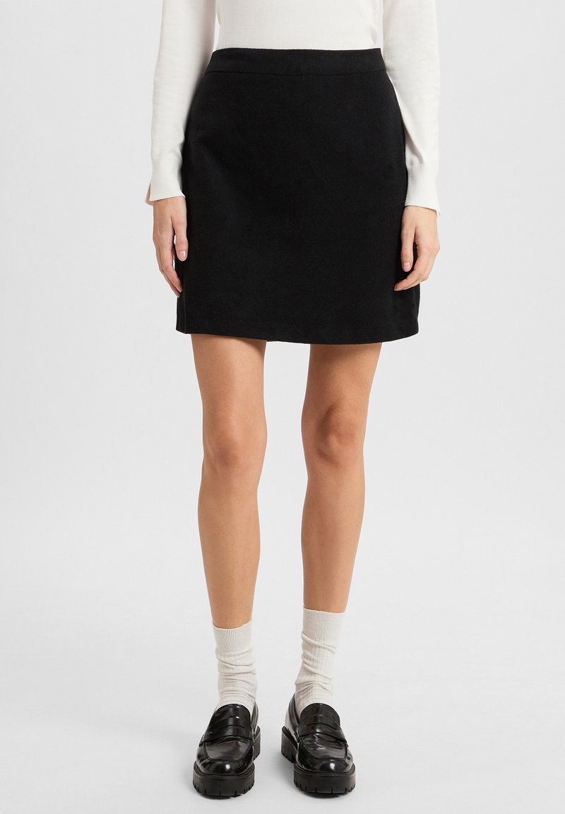 Schwarzer A-Linien-Rock aus glattem Stoff, mit einem eng anliegendem Bund und einem einfachen, sauberen Saum. Kombiniert mit klobigen schwarzen Loafers und hellen Socken.