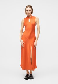 Mujer con vestido midi sin mangas color naranja, con escote tipo ojo de cerradura y abertura alta en la pierna, combinado con zapatos negros sin cordones.