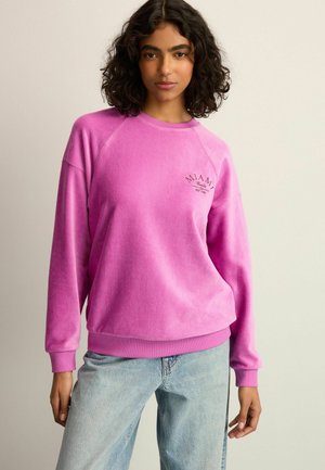 Rosa Sweatshirt mit strukturiertem Finish, Raglanärmeln und einem grafischen "MIAMI"-Design auf der Brust. Kombiniert mit hellblauen Jeans.