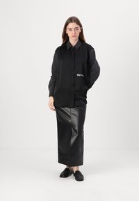 Melna pogu krekla ar kontrastējošām tumšām piedurknēm un krūšu logo, saskaņota ar garu melnu mākslīgās ādas svārku un melnām kurpēm.