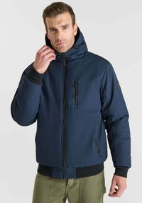 Giacca isolante blu navy con cappuccio, presenta una zip frontale, tasca sul petto con zip e polsini a coste per maggiore calore.