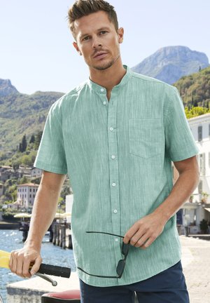 Camisa de hombre de manga corta y cuello en verde claro, de tela texturizada con un bolsillo frontal, con botones blancos y un corte relajado.