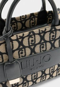Handtasche mit einem gemusterten, beigen Baumwollkörper, der schwarze Logos zeigt, ergänzt durch schwarze Lederakzente und einen Reißverschluss.