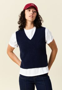 Marinblå stickad väst med V-ringning, lager på lager över en vit t-shirt. Har en ribbad nederkant och ett mjukt, strukturerat tyg.