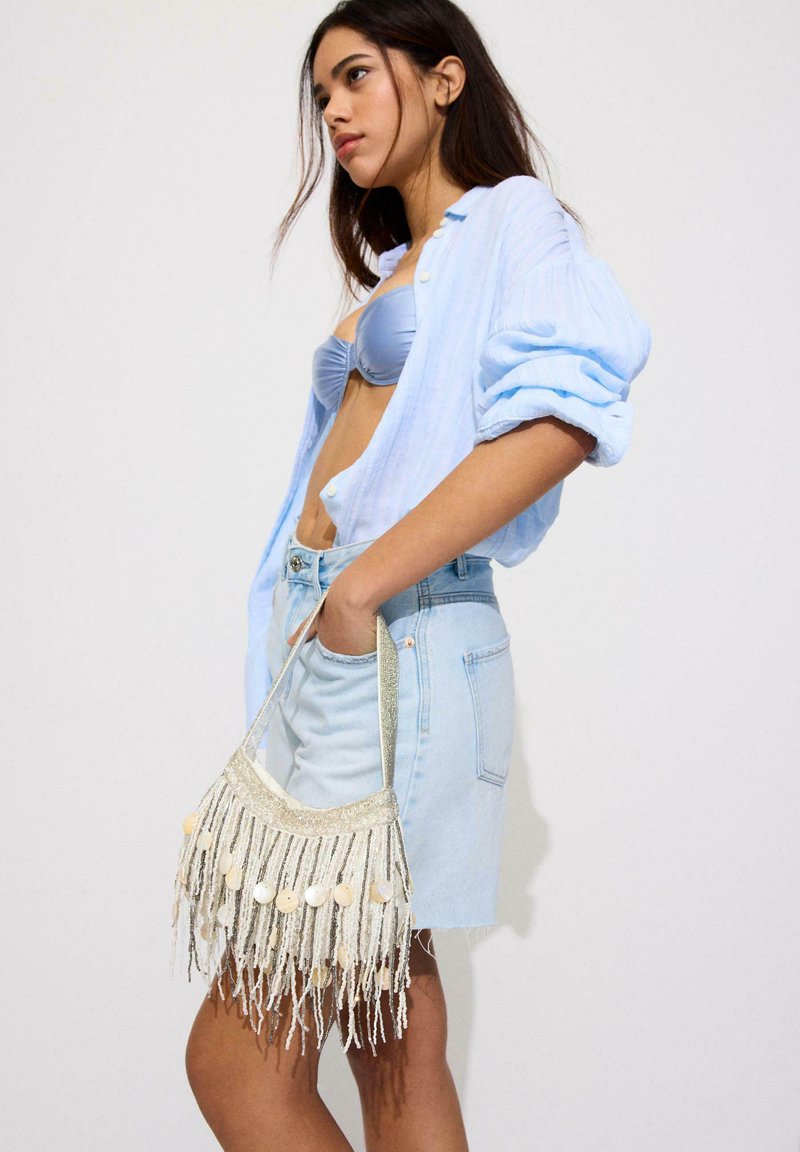 Jeune femme portant une chemise bleu clair déboutonnée sur un soutien-gorge assorti, jupe en jean, tenant un sac à main à franges perlées sur le côté.
