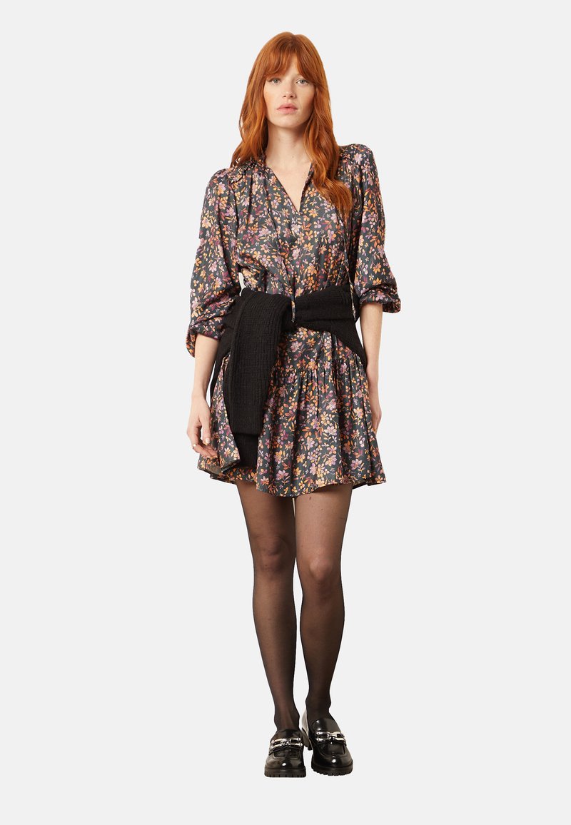 Vrouw met rood haar die een bloemenjurk draagt, zwarte panty, een zwarte trui om haar taille geknoopt, en zwarte loafers met zilveren gespen.