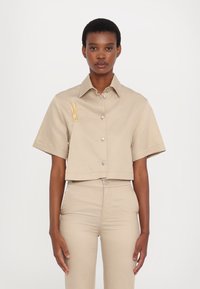 Beige cropped shirt with short sleeves, front button closure, and a small embroidered detail. Made from a smooth, lightweight fabric.