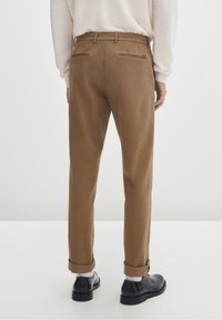 Mężczyzna w podwiniętych brązowych spodniach, białych skarpetach, czarnych butach i jasnym swetrze z długim rękawem, pokazany z tyłu.