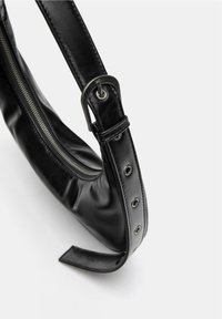 Fekete bőr kézitáska ívelt formával, cipzáros záródással és fém kiegészítőkkel. A pánt gombos részleteket és állítható csatot tartalmaz.