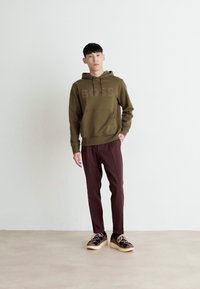 Olivgrüner Kapuzenpullover mit Fronttasche und "BOSS"-Logo, kombiniert mit weinroten, schmal zulaufenden Hosen und schwarzen Sneakern mit hellen Sohlen.