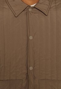 Quiltad tygjacka i brunt med spetsig krage och tryckknappar. Har synlig sömnad och två framfickor.