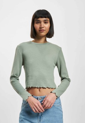 Ung kvinne med rett svart bob-frisyre iført en lys grønn kortermet topp med lange ermer og høyt liv blå denimjeans.