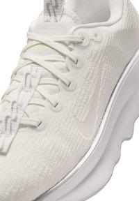 Baskets de sport blanches avec tige en tissu texturé, lacets allongés, semelle flexible et accents discrets en gris sur le côté.