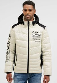 Man wearing a white and black padded jacket with hood, zipper front, printed text on chest and sleeve, and blue jeans.