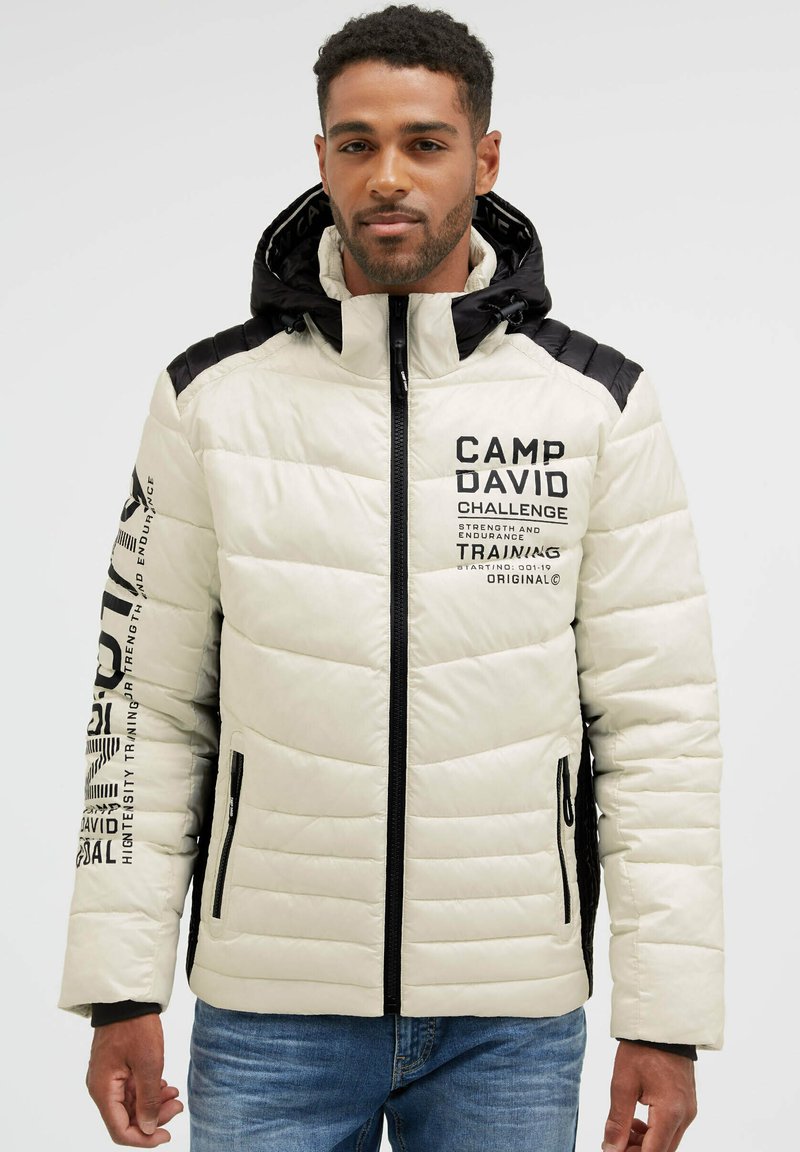 Man wearing a white and black padded jacket with hood, zipper front, printed text on chest and sleeve, and blue jeans.