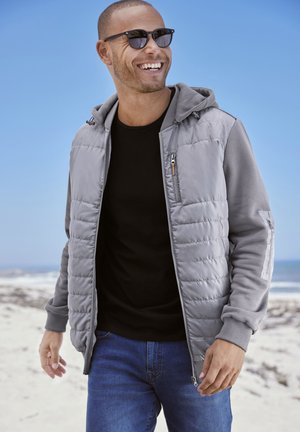 Gray padded jacket with a hood and contrasting gray sleeves, paired with a black t-shirt and blue jeans, set against a beach background.