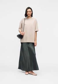 T-shirt oversized beige, jupe maxi en satin vert foncé et ballerines roses. Le mannequin porte un sac à bandoulière noir texturé. Fond neutre.