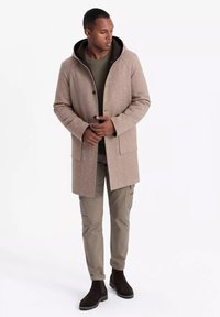 Abrigo de mezcla de lana beige con capucha, que cuenta con dos bolsillos frontales y un cierre de botón. Combinado con una blusa verde oliva, pantalones cargo beige y botas marrones.