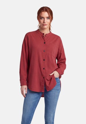 Frau mit roten Haaren, die ein rostrot geknöpftes Langarmhemd und blaue Jeans trägt und mit einer Hand in der Tasche steht.