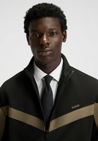 Ung mand med kort krøllet hår iført en sort zip-up jakke med beige striber over en hvid skjorte og struktureret sort slips på en hvid baggrund.