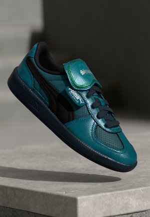 Zapatilla deportiva en verde azulado y negro con acabado brillante, collar acolchado, inserciones de malla y una correa de velcro. Suela de goma con patrones de agarre texturizados.
