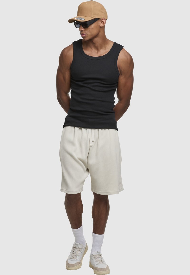 Sort ribstrikket tanktop, cremefarvede teksturerede shorts, beige kasket, mørke solbriller, hvide atletstrømper og lyse sneakers.