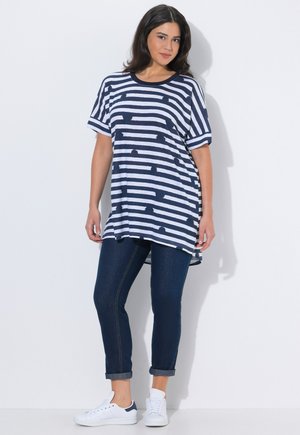 Donna che indossa una maglietta oversize a righe blu navy e bianche, jeans scuri risvoltati e sneakers bianche, in piedi su uno sfondo bianco semplice.