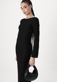 Vestido preto de manga longa com padrão texturizado de ondas. O modelo segura uma pequena mala preta arredondada. O fundo liso realça o foco na roupa.