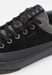 Zapatillas negras de ante y malla con una suela de goma texturizada, ojetes redondos y una correa de logo negra prominente en la lengüeta.