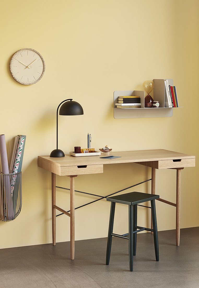Lichthouten bureau met twee lades, zwarte lamp en donkergroene kruk, tegen een gele muur. Heeft een wandplank en een klok.