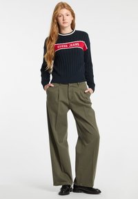 Marinblå kabelstickad tröja med vit och röd "GUESS JEANS" logotyp, kombinerad med högmidjade olivgröna vida byxor och svarta skor.