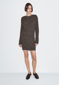 Dunkelbrauner, gestrickter Pullover und dazu passender Rock mit einem strukturierten Zopfmuster, rundem Ausschnitt und langen Ärmeln. Kombiniert mit schwarzen Loafers.